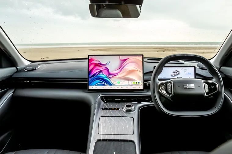 Interior of a Geely vehicle showing a large landscape-oriented touchscreen infotainment display, a secondary driver-facing digital screen on a flat-bottomed steering wheel, and a minimalist dark dashboard with silver panel detailing, with a beach visible through the windscreen
