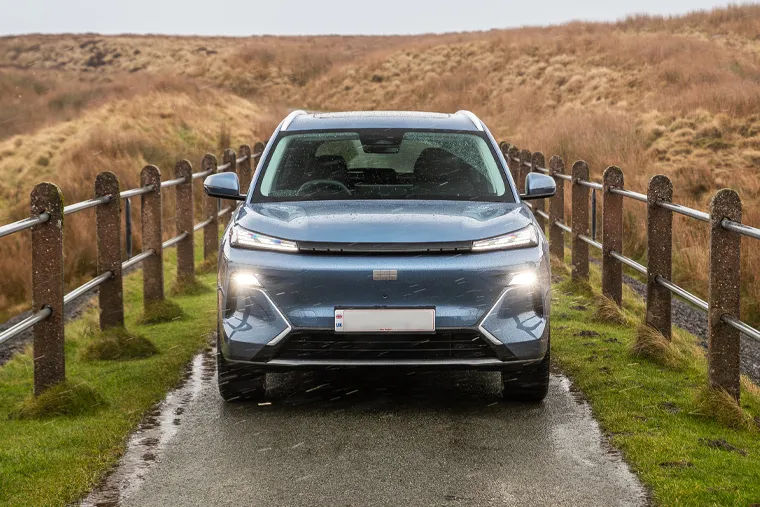 Head-on view of a Geely SUV in blue driving along a narrow rural track lined with wooden fencing, headlights illuminated in rainy conditions