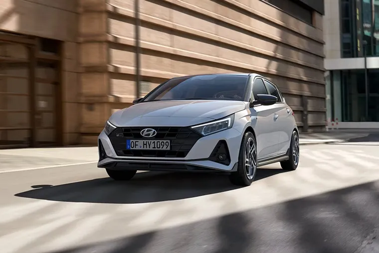 White Hyundai hatchback driving through a modern urban setting, showcasing sleek and dynamic design.