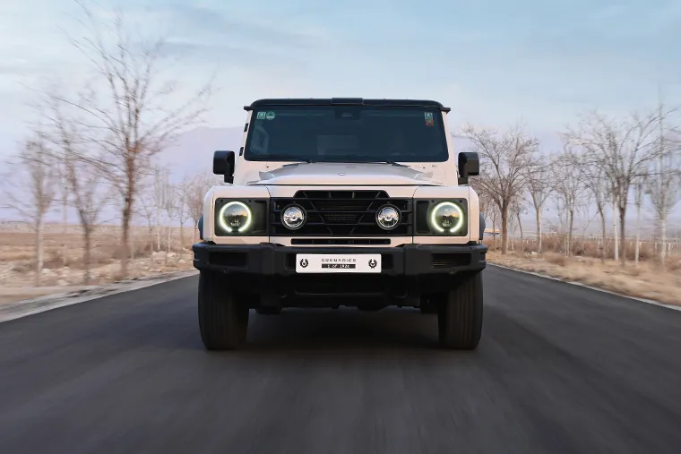Front‑view INEOS Grenadier SUV driving on rural road, displaying distinctive round headlights, robust bumper and purpose‑built off‑road styling.