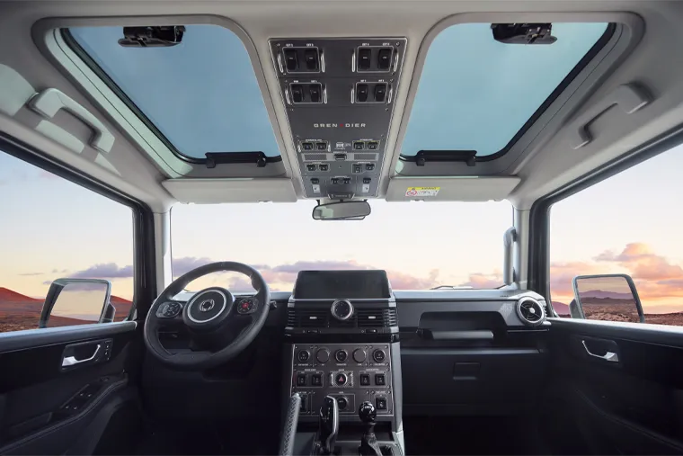 INEOS Grenadier interior view showing rugged dashboard, overhead control panel and dual sunroofs with panoramic sky, highlighting the off‑road SUV’s functional cockpit design.