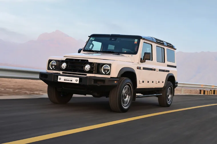 INEOS Grenadier white 4x4 SUV driving on open road with mountain backdrop, showcasing its rugged off‑road capability and premium adventure design.