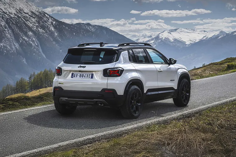Rear view of white Jeep Avenger SUV driving on a scenic mountain road with snow-capped peaks in the background.