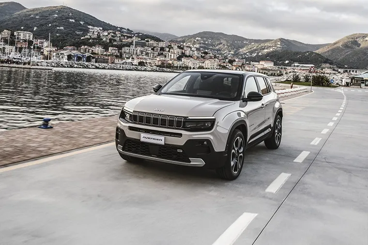 Jeep Avenger SUV driving on a coastal promenade beside a marina and hillside townscape.