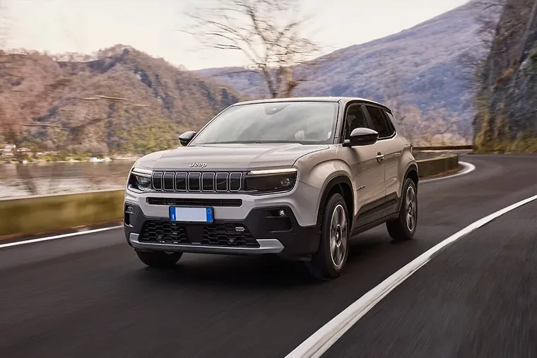 Front view of Jeep Avenger SUV driving on a winding mountain road beside a lake in early spring.