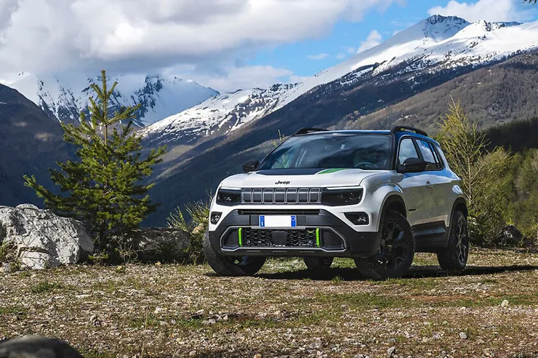 White Jeep SUV parked on a rocky mountain trail with snowy peaks and pine trees in the background.