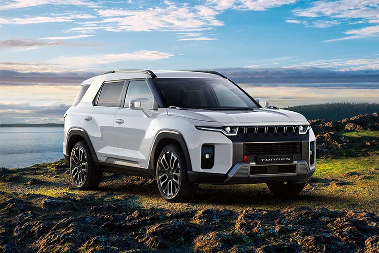 White KGM Torres SUV parked on a rocky landscape beside a lake under a bright blue sky with clouds at sunset