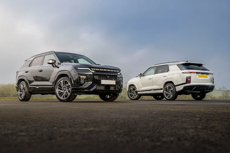 Front and rear view of two 2023 KGM Torres SUVs parked on an open road, with a black model on the left showing the front fascia and a white model on the right showing the rear design, under a cloudy sky.