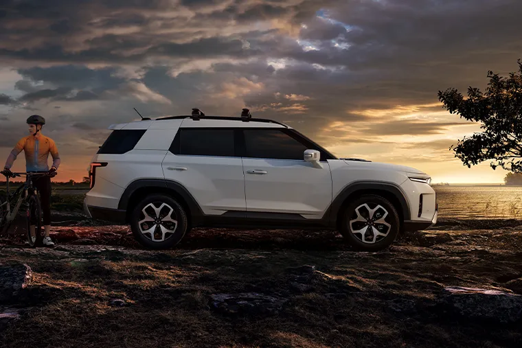 White KGM Torres SUV parked beside two cyclists at sunset, with dramatic clouds and a lone tree in the background