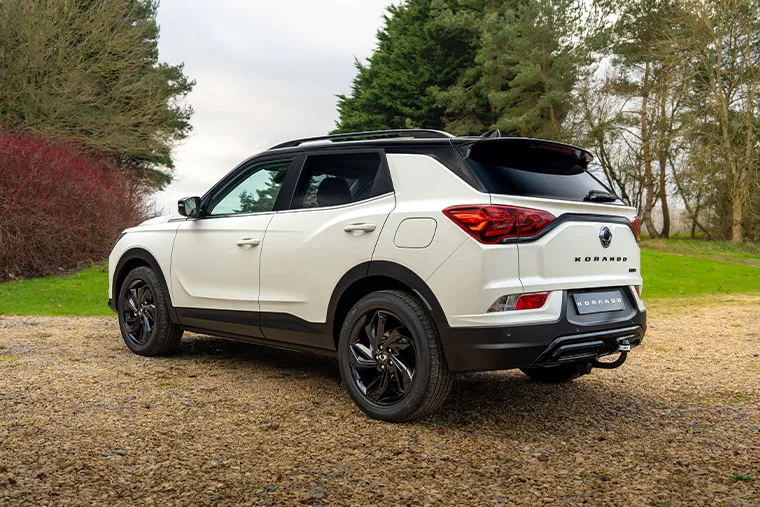 Rear three-quarter view of white KGM Korando SUV parked on a gravel driveway surrounded by trees