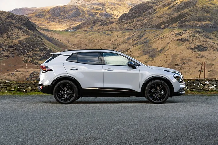 Side profile of a white Kia Sportage SUV parked in front of scenic mountain landscape.