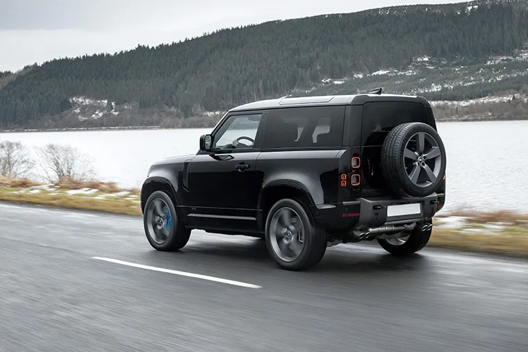 Black Land Rover Defender 90 driving on a lakeside road with forested hills in the background.