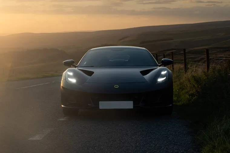 Black Lotus Emira sports car front view with headlights on, driving on a country road at sunset