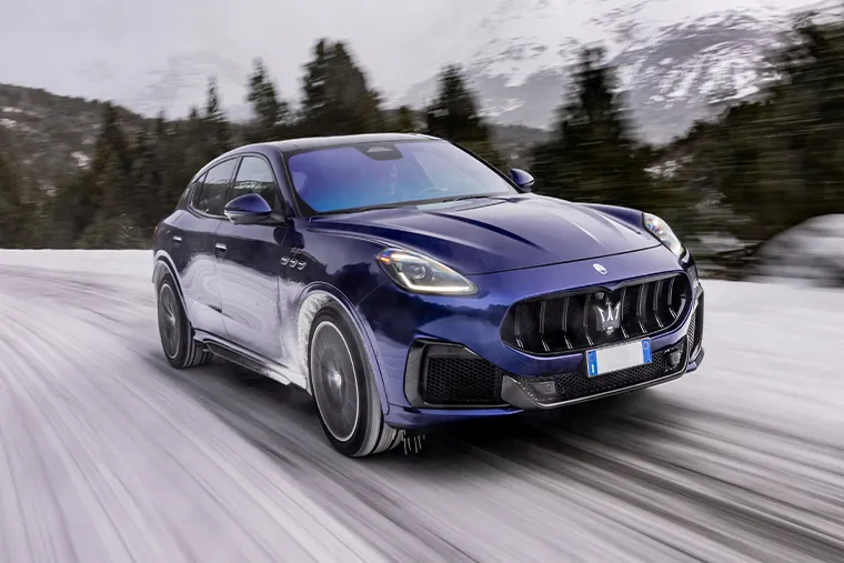 Blue Maserati Grecale SUV driving in snowy mountain landscape, rear three-quarter view