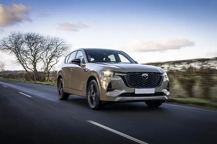 A beige Mazda SUV driving on a rural road with blurred hedgerows and a wintery tree in the background.