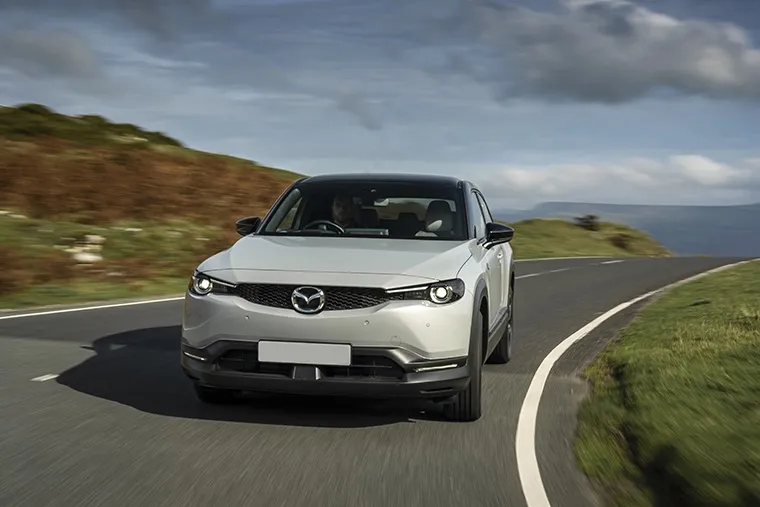 A silver Mazda MX-30 electric SUV driving along a winding country road with dramatic hills and sky in the background.