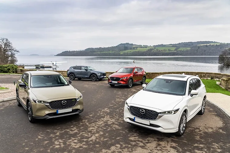 A lineup of Mazda SUVs, including models in gold, white, red, and grey, parked near a scenic lakeside with hills in the background, showcasing the variety in the Mazda SUV range.