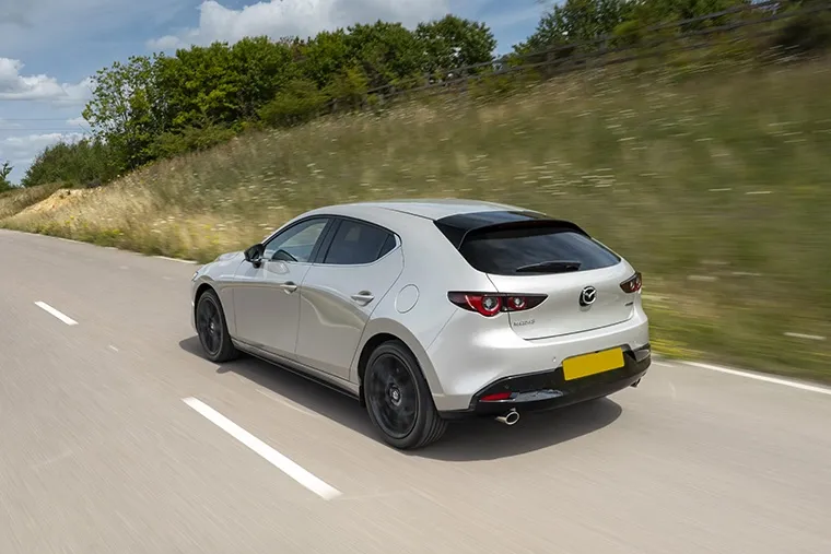 A silver Mazda3 hatchback driving along a countryside road on a sunny day