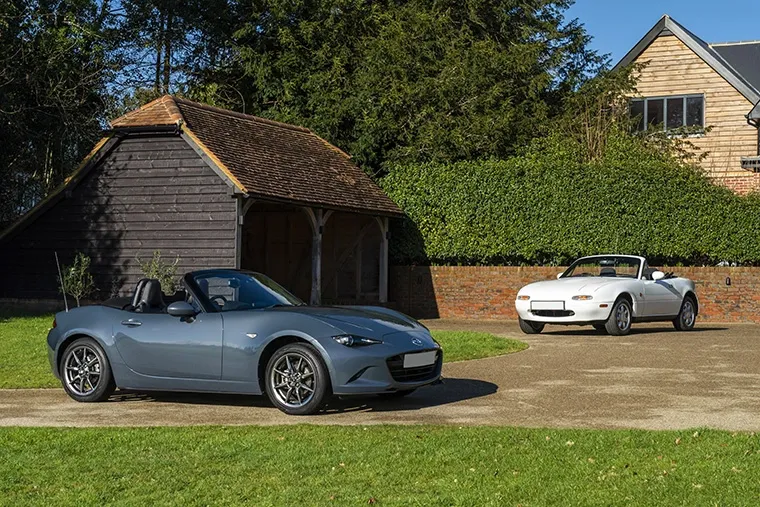 Two generations of the Mazda MX-5 parked in front of a wooden carport and brick house.