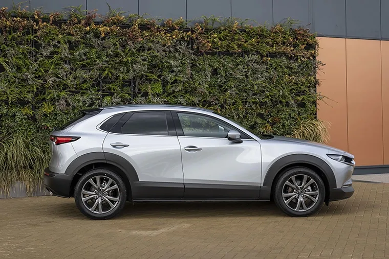 Side profile of a silver Mazda CX-30 SUV parked in front of a modern building with a green wall, showcasing its stylish and practical design.