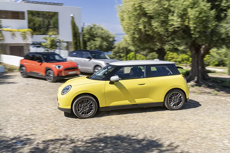 Yellow MINI Electric driving on a sunny day near modern home with two parked MINI SUVs.