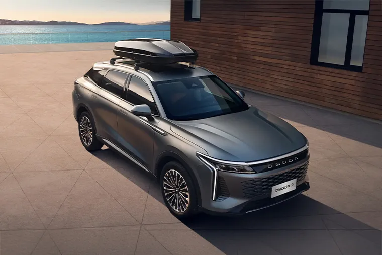Silver Omoda 9 SUV parked by a modern wooden building near the sea, featuring a roof box and sleek aerodynamic styling.