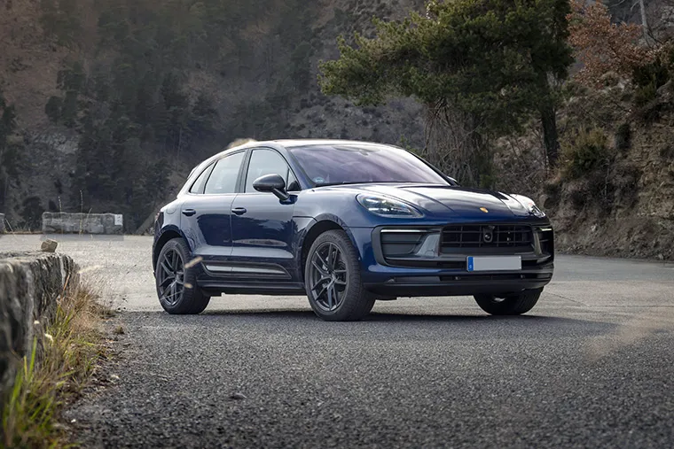 Dark blue Porsche Macan SUV parked on a mountain road with forested hills in the background at sunset.
