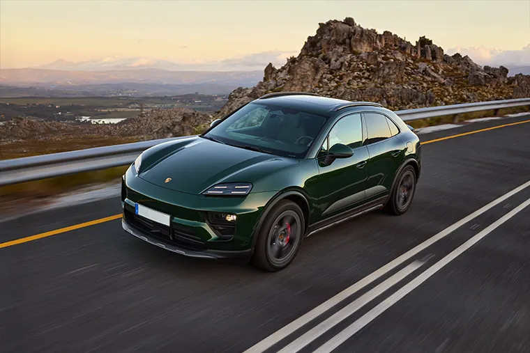 Dark green electric Porsche Macan driving on a mountain road at sunset.