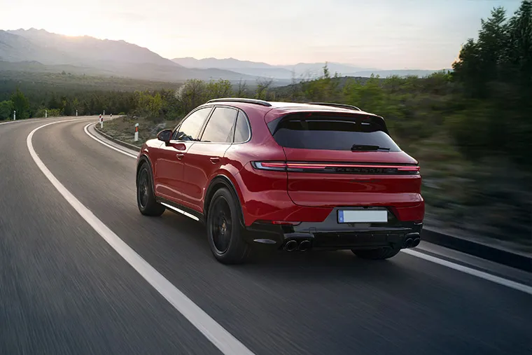 Rear view of a red Porsche Cayenne SUV driving on a winding mountain road at sunset.