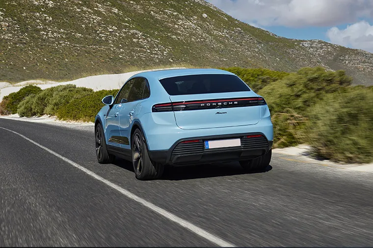 Light blue Porsche Macan EV driving on a scenic coastal road with green hills.