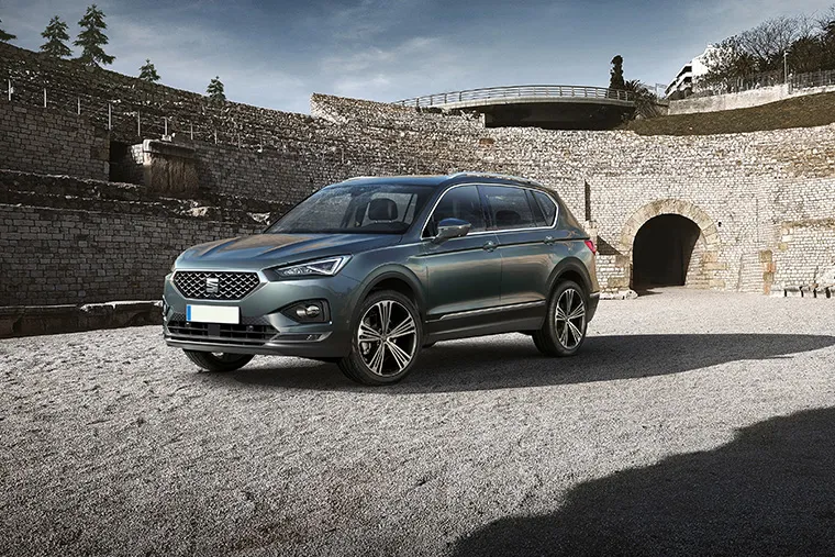 SEAT Tarraco SUV parked in front of ancient stone ruins with modern buildings in the background under a blue sky.