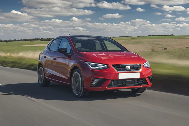 Red SEAT Ibiza hatchback driving through open countryside under a bright blue sky with scattered clouds.