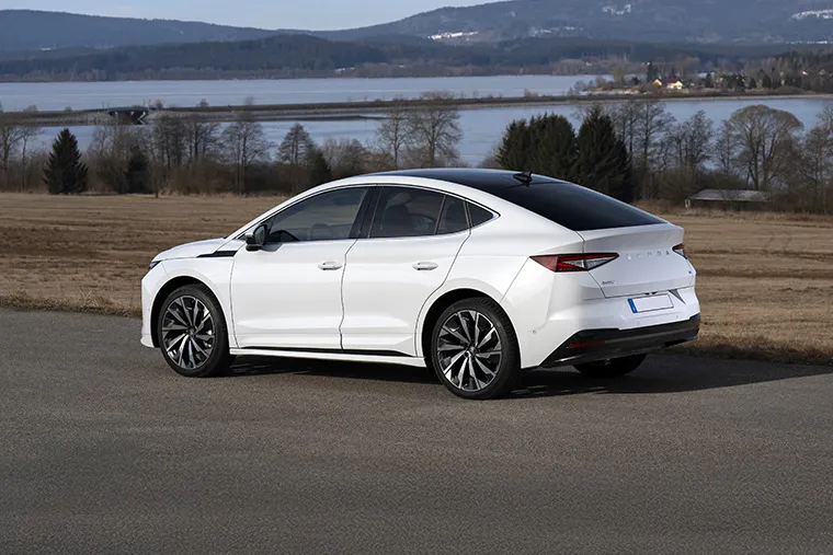 Rear-side view of a white Skoda Enyaq Coupé parked on a countryside road with a scenic lake.