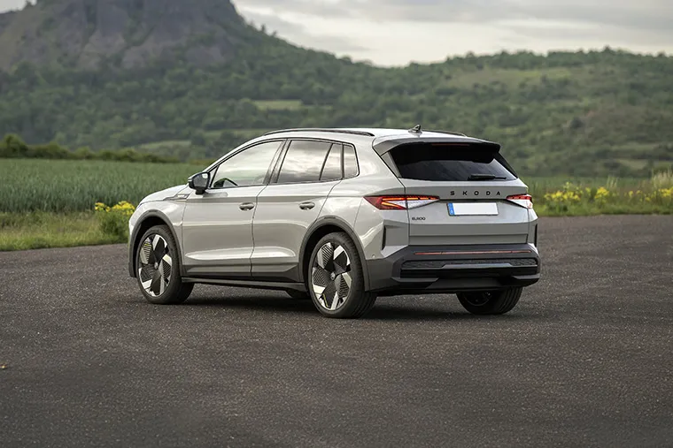 Rear-side view of a silver Skoda Enyaq electric SUV parked in front of green hills and a rocky mountain landscape under a cloudy sky.