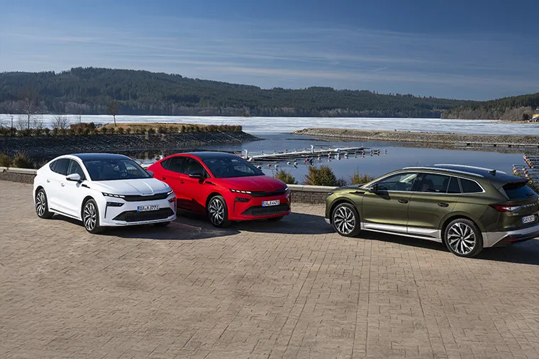 Three Skoda electric vehicles in white, red, and green parked by a scenic lakeside marina with forested hills in the background.