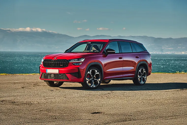 Front-side view of a red Skoda Kodiaq SUV parked near the ocean with mountains in the background on a clear sunny day.