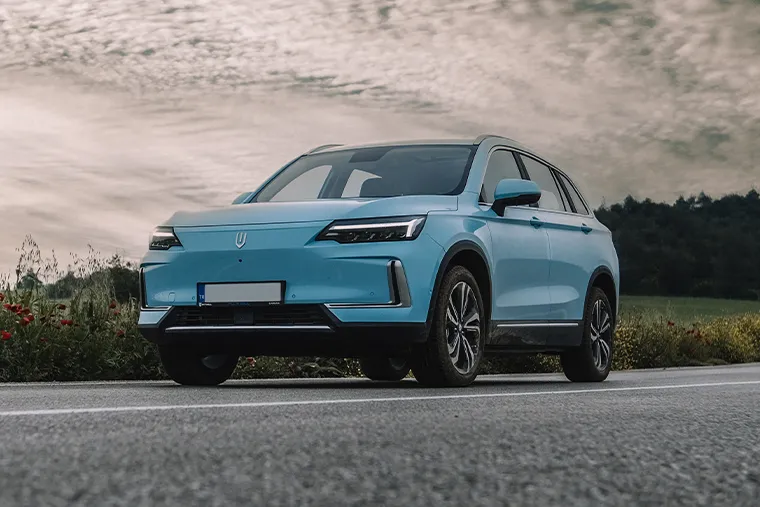 Light blue Skywell ET5 electric SUV parked on a scenic country road beside wildflowers, with a dramatic cloudy sky and rolling hills in the background.