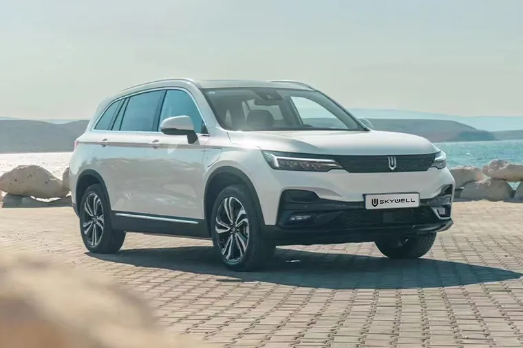 White Skywell ET5 electric SUV parked on a sunlit coastal promenade with the sea and distant hills in the background.