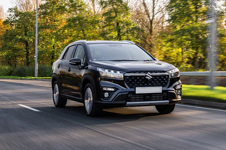Front view of 2025 Suzuki S-Cross in black driving on suburban road with autumn trees in background.