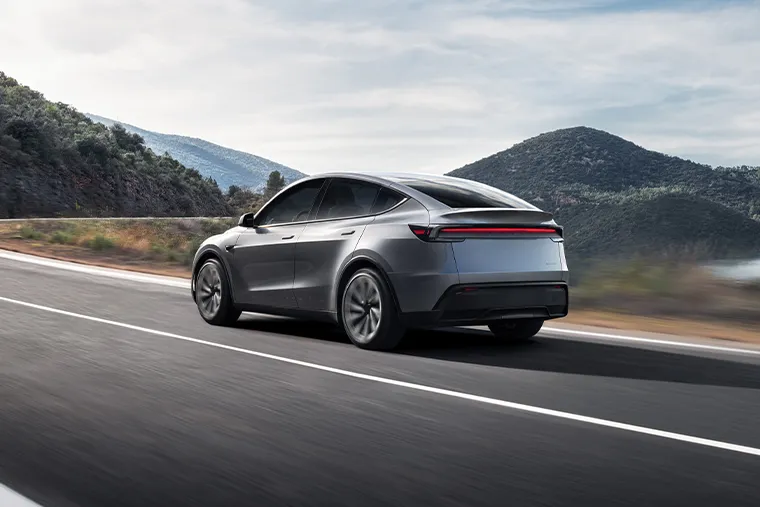Silver electric saloon driving along a scenic mountain road beside a lake under a clear blue sky.