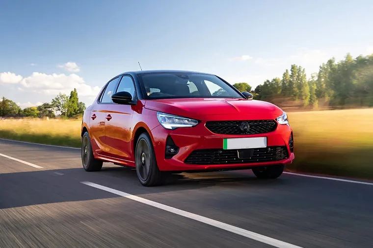 Red Vauxhall Corsa driving on a countryside road under a clear blue sky.