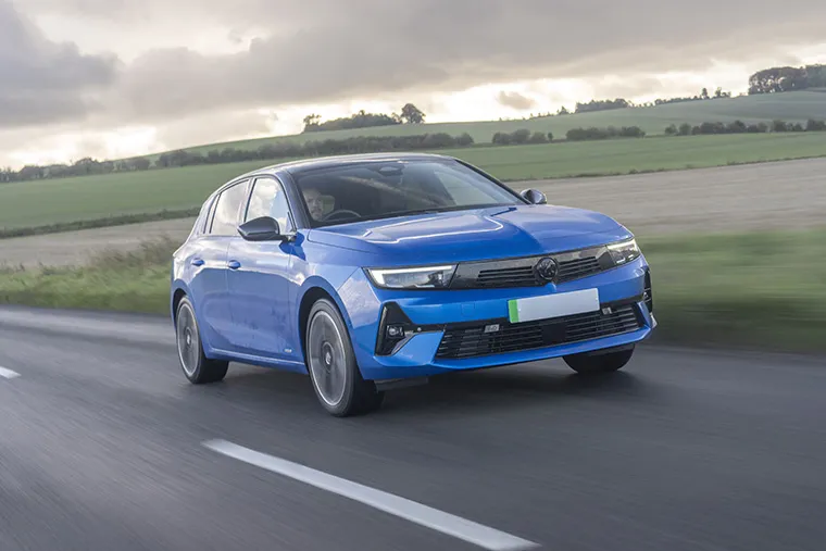 ront view of a blue Vauxhall Astra hatchback driving on a country road with green fields and a cloudy sky in the background.